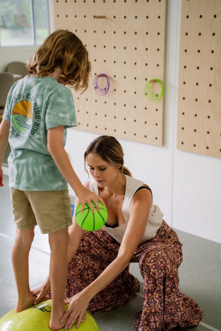 A paediatric physiotherapist and a child standing on a ball in a therapy room.